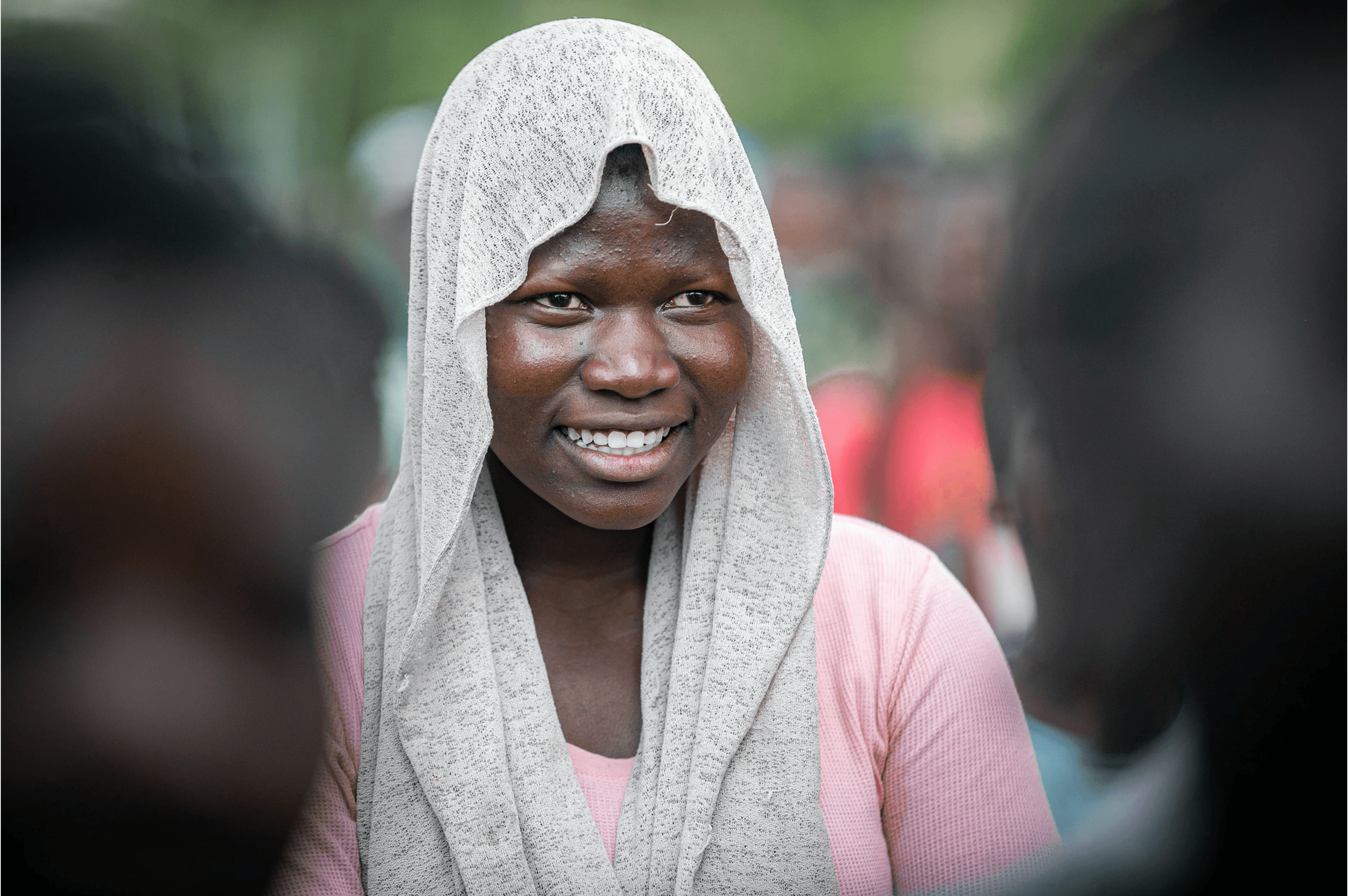 A young woman wrapped in a grey woven headscarf smiles candidly, standing among a softly blurred crowd.